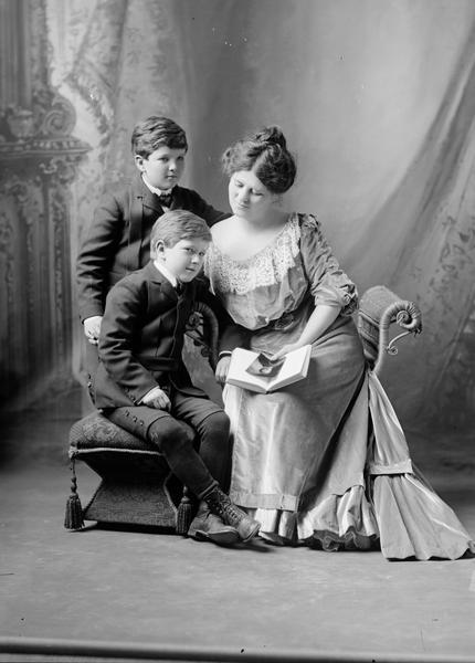 Formal seated portrait of Belle Case La Follette, the wife of Robert M. La Follette, Sr., and her two sons, Robert M. La Follette, Jr., (standing) and Philip Fox La Follette. Mrs. La Follette is holding a photograph of her husband.  Her dress is one that she would have worn as the wife of the governor on formal occasions.