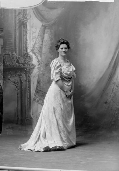 Formal, full-length portrait in front of a painted backdrop of Belle Case La Follette, the wife of Robert M. La Follette, Sr. Although the negative jacket identified the portrait as 1903-1904, the La Follette Family album, which was assembled by Isabel La Follette, identifies it as 1905-1906, which seems more likely. It is also likely the dress is a new one purchased to wear in Washington, D.C.