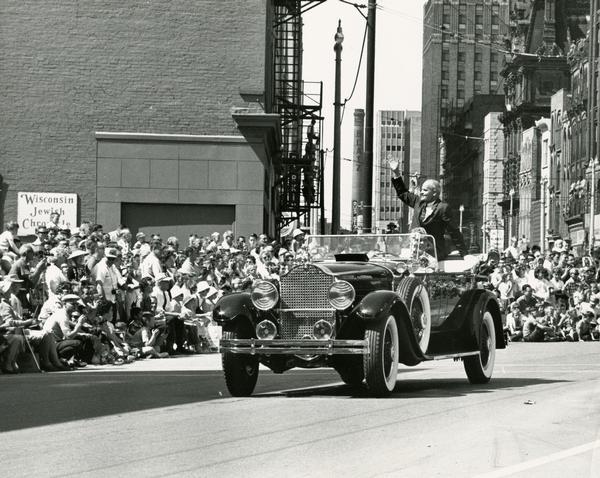 Senator William Proxmire waves to the crowd from an open antique automobile during a parade in downtown Milwaukee.