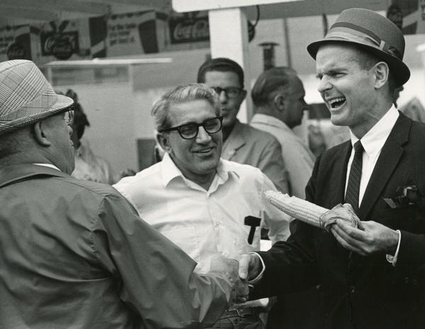 Senator William Proxmire shakes hands with constituents at the Wisconsin State Fair while eating an ear of corn.