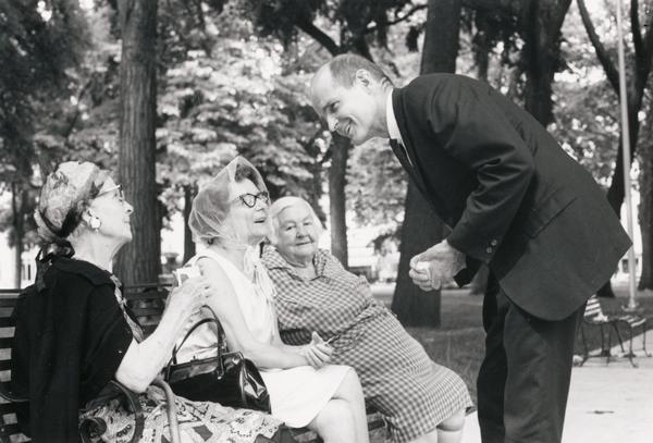 Senator William Proxmire greets three elderly women seated on a bench at the Capitol Square.