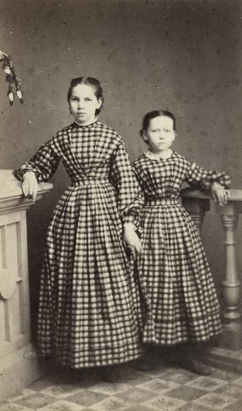 Formal studio portrait of Charlotte (left), and Augusta Lueders of Sauk City, each sister wearing dresses made from the same plaid fabric.  Their father was E.G.J. Lueders who came to Sauk City in 1851.