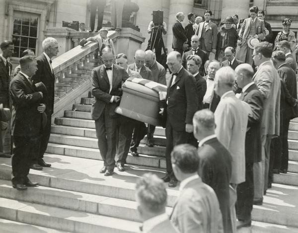 Pallbearers (who include Senator Herman Ekern), carry the flag draped casket of Senator Robert M. La Follette, Sr., down the steps of the Wisconsin Capitol to the waiting hearse.  In the background are Robert M. La Follette, Jr., Belle Case La Follette, and Philip Fox La Follette.