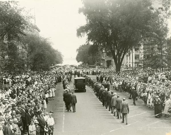 The automobile bearing the casket of Senator Robert M. La Follette, Sr., begins it journey from the Wisconsin Capitol to Forest Hill Cemetery.  Here it is about to leave the Capitol grounds at Monona Avenue before turning left onto Main Street.  The vehicle is surrounded by an enormous crowd of citizens.
