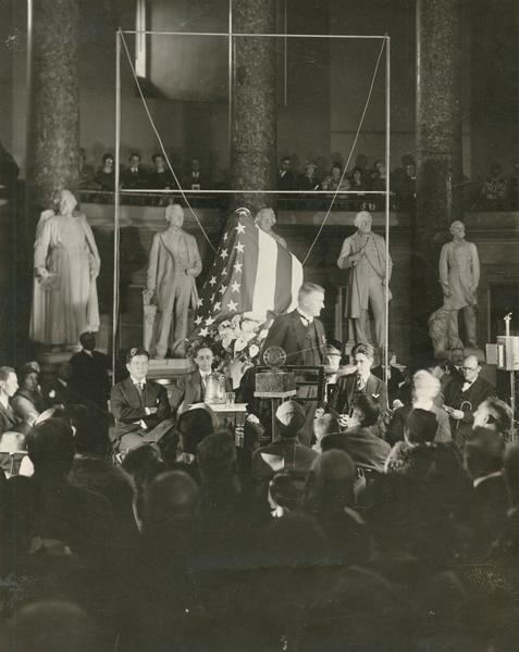 Ceremony for the unveiling of the statue of Robert M. La Follette, Sr., in Statuary Hall in the U.S. Capitol. On the platform, left to right, are Senator Robert M. La Follette, Jr., an unidentified Baltimore newspaperman, Senator John J. Blaine of Wisconsin at the podium, and Philip Fox La Follette. Behind them is the framework that will be usued to unveil the flag-draped statue sculpted by Jo Davidson, as well as several statues.