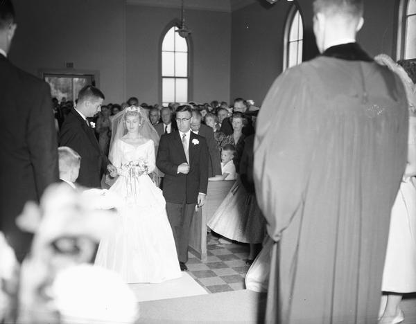 The groom offers his arm, while the father of the bridge prepares to step away at the Saalzaa-Ziemer wedding at the Argyle Methodist Church. Watching are the wedding guests in the background and the minister (believed to be Kenneth McMurtrey) in the right foreground.