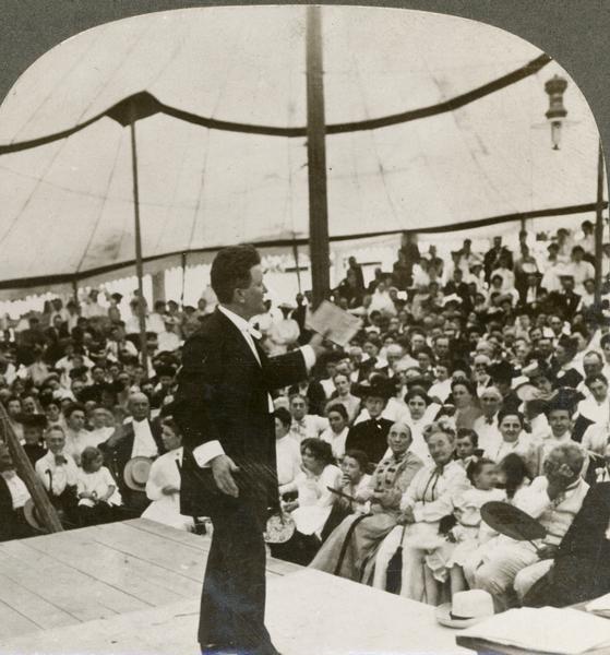 Senator Robert M. La Follette, Sr., speaking to an audience at Decatur, Illinois.  La Follette is standing on a raised platform inside a large tent.