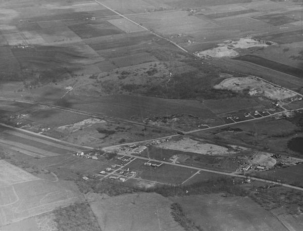 Aerial view, looking eastward from a point several miles outside the southwestern limits of Madison in the direction of Verona. Crossing the image diagonally from the left (north) edge to the lower right (south) corner is the Verona Road (Hwys. 18-151). Existence of an important mineral resource is evidenced in the photo by the three installations along the right side of the photo. The lower building is the plant of paving contractors Rein & Schultz. The middle building is the gravel pit and plant of Hammersley Stone Co. and the upper, the pit of the Wingra Stone Company.