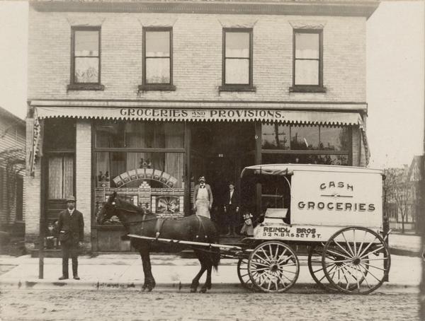A horse and wagon and men are posing in front of Reindl Brothers Groceries and Provisions store at 32 North Bassett Street. Text on the wagon reads: "Cash. Groceries."