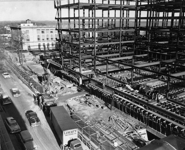Elevated view of the City County Building under construction. Automobiles are parked in the street and a truck with a semi-trailer is parked in front of a cement truck.
