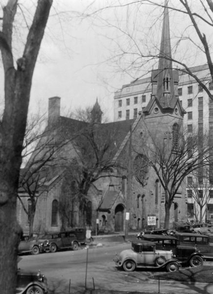 View from across the street towards the Congregational Church at the corner of North Fairchild Street and West Washington Avenue. To the left of the church is the Gates of Heaven Synagogue. Automobiles are parked on both sides of the street, and in the background is the Wisconsin Power and Light Company.