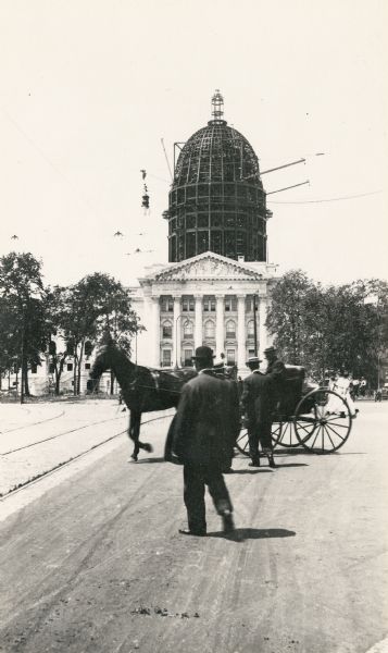 Wisconsin State Capitol under construction, with men and a horse-drawn buggy in the foreground.