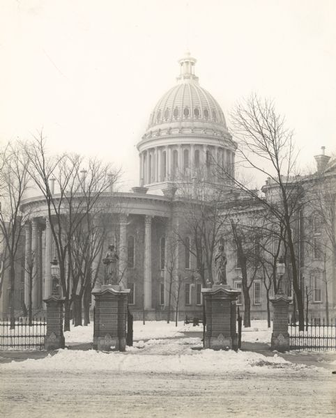 View from street through gates towards the Third Wisconsin State Capitol, the second in Madison, as seen from East Washington Avenue. The iron fence and stone gateways around the perimeter of the Capitol Park were designed by Stephen V. Shipman, who had also designed the dome, completed in 1873. There are iron sculptures on top of the columns.