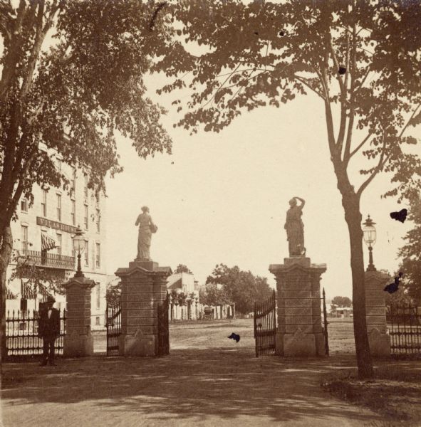 View of the gate to the Wisconsin State Capitol grounds, looking out to the street. On the left on the opposite side of the street is a building with a sign that reads: "Vilas Hotel."