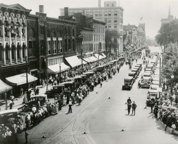 The funeral procession of Senator Robert M. La Follette, Sr., begins its journey from the Capitol to Forest Hill Cemetery, travelling  east around the Square on Main Street.  The street is crowded with mourners and automobiles.