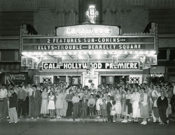A crowd is standing on the sidewalk in front of the Eastwood Theater. A sign behind them reads: "Gala Hollywood Premiere." The "Premiere" is of thirty local people impersonating movie stars. The marquee reads: "2 Seatures Sun-Cohens and Kellys in 'Trouble' and 'Berkeley Square.'"