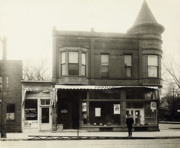 Ehrman's Delicatessen, located at 300 State Street, on the corner of State and Henry Streets. One man is standing on the sidewalk, and another man is standing inside the store. The sign on the door on the left side of the building reads: "J.J. Schrodt Chiropractor Spinal Adjustments". A small storefront on the left has a sign that reads: "Olympia Shoe Shining Parlor, Chairs for Ladies and Gents, Hats Cleaned".