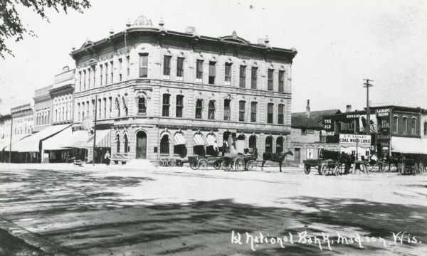 The First National Bank, on the corner of North Pinckney and East Washington on the Capitol Square. Horse-drawn carriages are on East Washington Avenue.