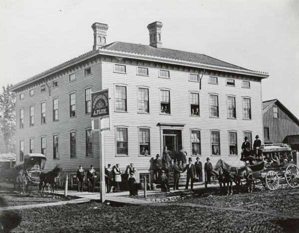 The Flom Hotel or American House, 323 East Main Street, at the corner of South Hancock Street. The Vilas House, whose conveyance stands in front, was a rival establishment. The Andres Flom, proprietor of the Flom Hotel (wearing a silk top hat) shakes hands with a newly arrived guest (also in top hat, with bag). Other people include Flom porters, barkeeps, chambermaids and other members of the staff.