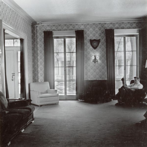 An interior view of the living room toward the front of 524 North Henry Street. A man is sitting in a chair on the right near the windows.
