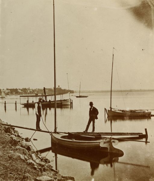 Two men pose on a boat docked at the Askew Steamboat Landing on Lake Monona off South Carroll Street. The old Governor Harvey residence is in the background on the far shoreline.