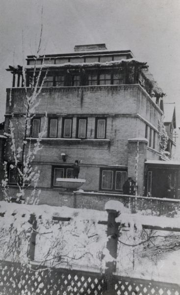 The Robert Lamp house, 22 North Butler Street, designed by Frank Lloyd Wright, architect. A man wearing a coat and hat is standing at the front right corner of the building, and a child, also wearing a coat and hat, is standing in a large planter on top of a brick wall holding a shovel or broom over their shoulder.
