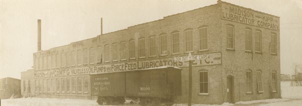 Exterior view of the Madison-Kipp Company during winter with a railroad car parked in front.