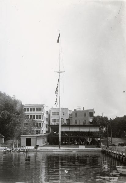 Mendota Yacht Club, later the site of James Madison Park. A mast with flags is standing outside of the club near the water.