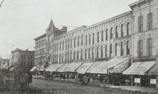 Pinckney Street.  The State Bank can be seen in the center, and to the right is a tailor, bookstore, and other retail shops.