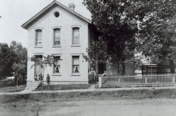 Dr. James A. Jackson residence, 123 North Pinckney Street. Dr. Jackson is standing by the fence; his wife Sydonia Jackson is in the center, lower window, a maid is in the other window. Daughter Alice is in the upper center window. On the steps, left to right are the children Joseph, Bettina and Russell with Reginald standing.