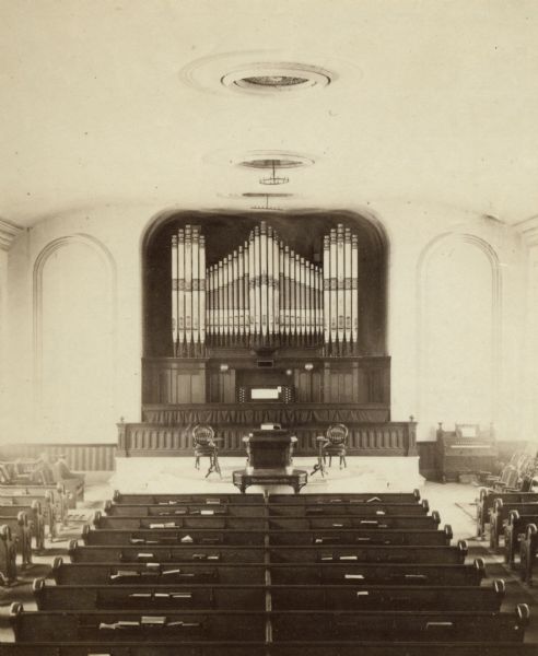 View of the interior of the Presbyterian church. There is an organ, piano and numerous pews visible.