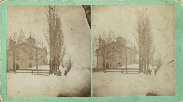 Stereograph of a wintery view of the Second Ward School House. A student stands at the edge of the fence line surrounding the school house.