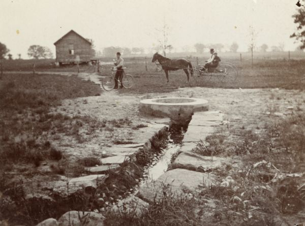 View of Silver Spring and the surrounding countryside. In the background, there is a man on a bicycle, two individuals in a horse-drawn vehicle and a small, wooden building.
