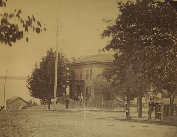 View of the Spooner residence on the south corner of Wilson and Carroll Streets, with the lake in the background. This was the home of John C., Phil H., and Roger Spooner. There are several women and men on the front porch, and a man on the right leans against a fence across the street from the house.