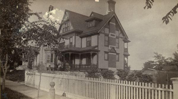 A side view of the Philip Spooner residence, 133 West Wilson Street. The family is gathered on the front porch of the house. There is a fence along the sidewalk in the foreground.