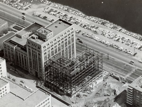 An aerial view of the construction of a new wing on a state office building. Automobiles are parked in a log along Lake Monona.