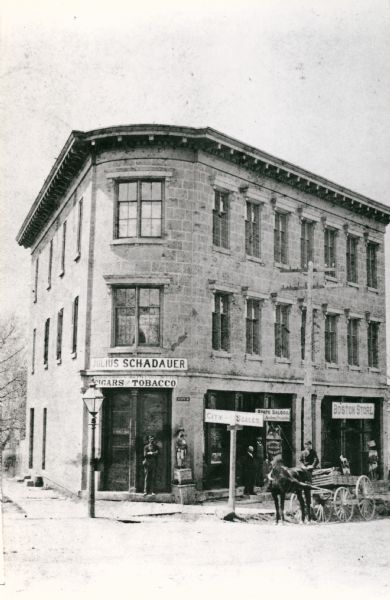View from street towards a man standing in front of the doorway to the Julius Schadauer's tobacco store on a street corner at 101 State Street. A man is on a horse-drawn wagon near the curb on the right. A sign at the street corner reads: "City-Scales." Signs on storefronts to the right of the tobacco store read: "State Saloon Anton Steinle." and "Boston Store."