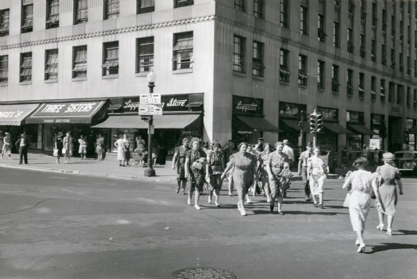 A group of pedestrians crossing the street outside of Liggetts Drugstore, located in the Tenney Building on Capitol Square.