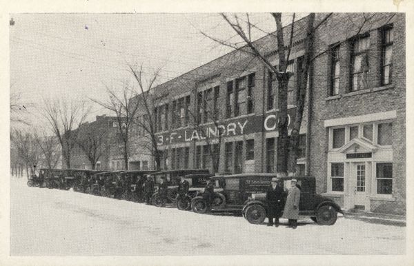 An exterior view of the 3-F Laundry Company located at 731 East Dayton Street. Two men are standing outside of the building next to a row of parked automobiles.
