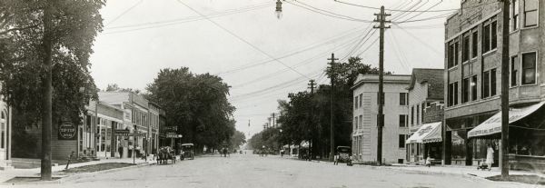 View down center of University Avenue with storefronts on both sides. The signs on the left read: "Hurley Bros. Grocers" and "Garage." On the right are signs for: "Motorcycles" and "Olwell Bros. Groceries."