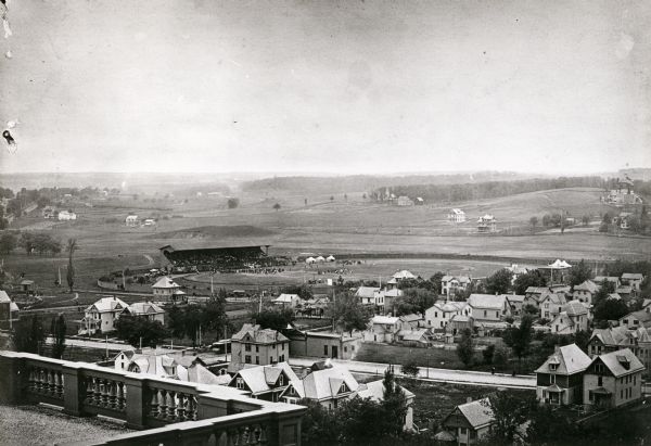 Elevated view of Camp Randall Field from the top of Bascom Hall, with University Heights in the background. A crowd is in the stands at the stadium, and a crowd is standing in the field. Tents are set up to the right of the stadium.