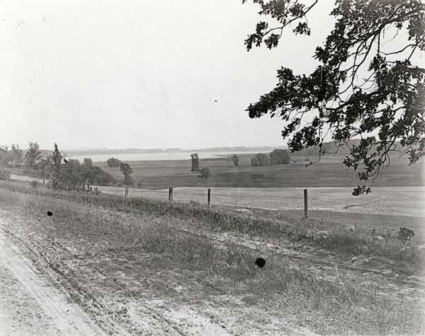 View from the western countryside towards Lake Wingra, looking east. In the foreground is a dirt road and fence.