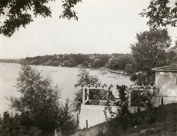 View along the steep shoreline of possibly Lake Mendota. In the foreground there is a veranda looking over the lake with various docks and boats in the background.