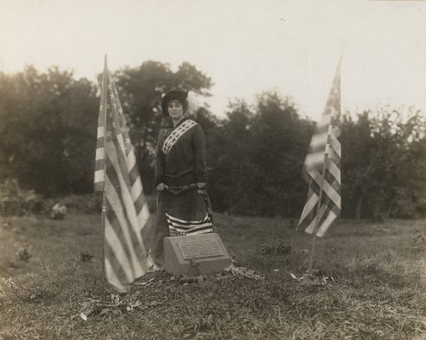 Sara (Mrs. Joseph) Mallon (Menominee) of Milwaukee, wearing a sash stands between two American flags behind a memorial that reads, "Indian Mounds: One of several groups of prehistoric burial. Linear and effigy mounds formerly located on the crest of the Monona-Wingra ridge. Several of these were surveyed by Increase A. Lapham in 1850. Village site was in the park below." This photograph was taken at the unveiling ceremonies of the memorial tablet which took place during the Fourth Annual Conference of the Society of American Indians at UW-Madison from October 6-11.
  
