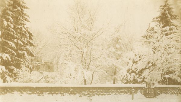 A snow-covered stone wall lines the perimeter of the Walker Castle grounds. Walker Castle is in the background behind trees. Located on East Gorham Street, the castle was torn down and part of the stones were used to construct the large stone residence at 137 East Gorham Street.
