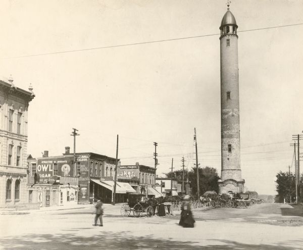 Water Tower located on East Washington Avenue. Horse-drawn wagons are parked in the center near the tower. Commercial buildings are in the background on the left.