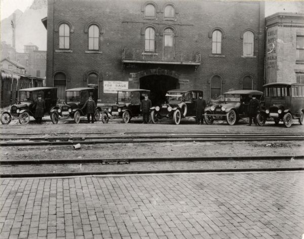Taxi cabs and their drivers are awaiting customers near the Chicago, Milwaukee, St. Paul and Pacific Railroad depot on East Wilson Street. In the center of the background at 643 East Wilson is the former residence and commercial building of William H. and Margaret Denison. They operated a "general teaming" business which specialized in "special piano moving," until about 1910. To the right is the former Eagle Hotel building, located at 631 East Wilson Street. Because of the railroad travelers, there were several hotels located in the vicinity.