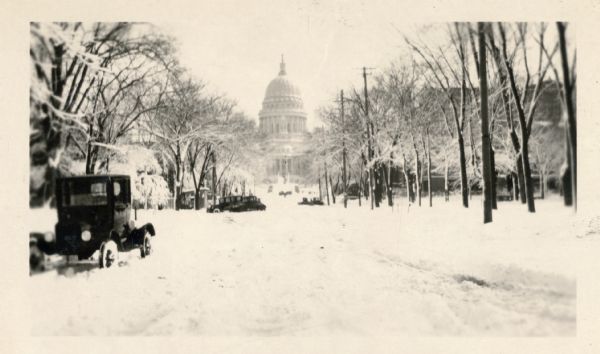 Winter scene of a deeply snow-covered West Washington Avenue, Madison, Wisconsin. In the background is the Wisconsin State Capitol building.
