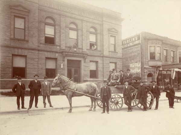 A group of men with a horse-drawn vehicle are standing outside of the Wisconsin Telephone Company Building, located at 16-18 S. Carroll Street.