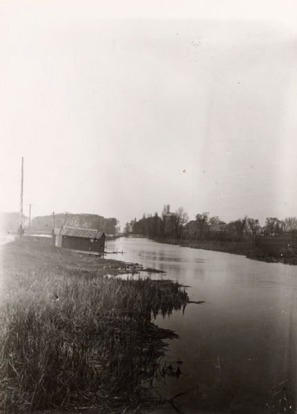 View along shoreline of the Yahara River, a small building along the shoreline near a road on the left. The Malt House is in the far background, prior to the Sherman Avenue improvement